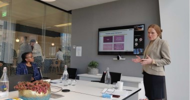 A woman stands in a suit jacket giving a presentation in a meeting room