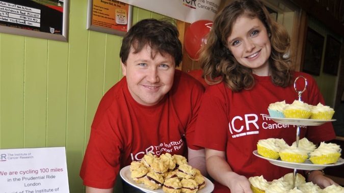 Bakesale fundraising - a man and woman wearing The Institute of Cancer Research branded red tshirts smile behind plates of cakes