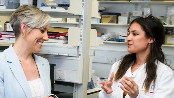 Corporate donors lab tour - Two women stand talking in a ICR laboratory, one has long dark hair and a lab coat and the other has short blonde hair and is wearing a blue suit