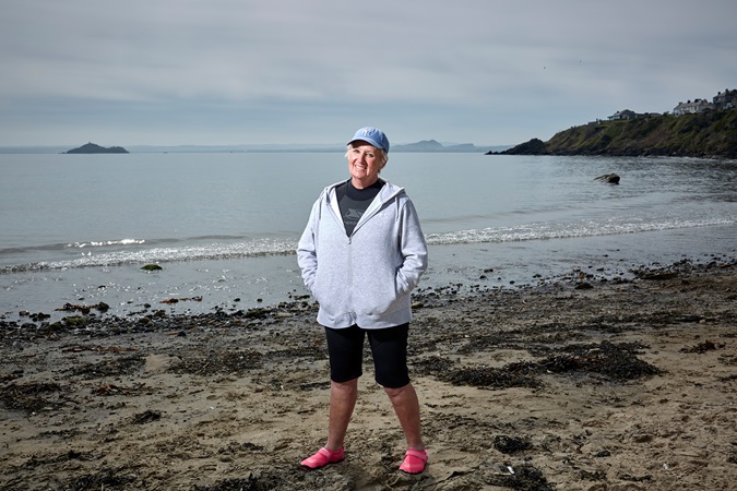 Karen- a woman wearing a blue cap and a wet suit with a grey hoodie over the top stands on a beach smiling at the camera