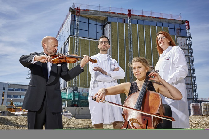 Philanthropy - unfinished symphony - Two scientists stand in lab coats in front of a half constructed building, in front of them a man in a suit stands playing a violin and a woman wearing a black dress sits playing a cello