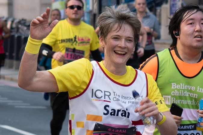 A woman holding a water bottle runs in a marathon, wearing a yellow and white shirt with "Sarah" printed on it. Other runners are visible in the background.