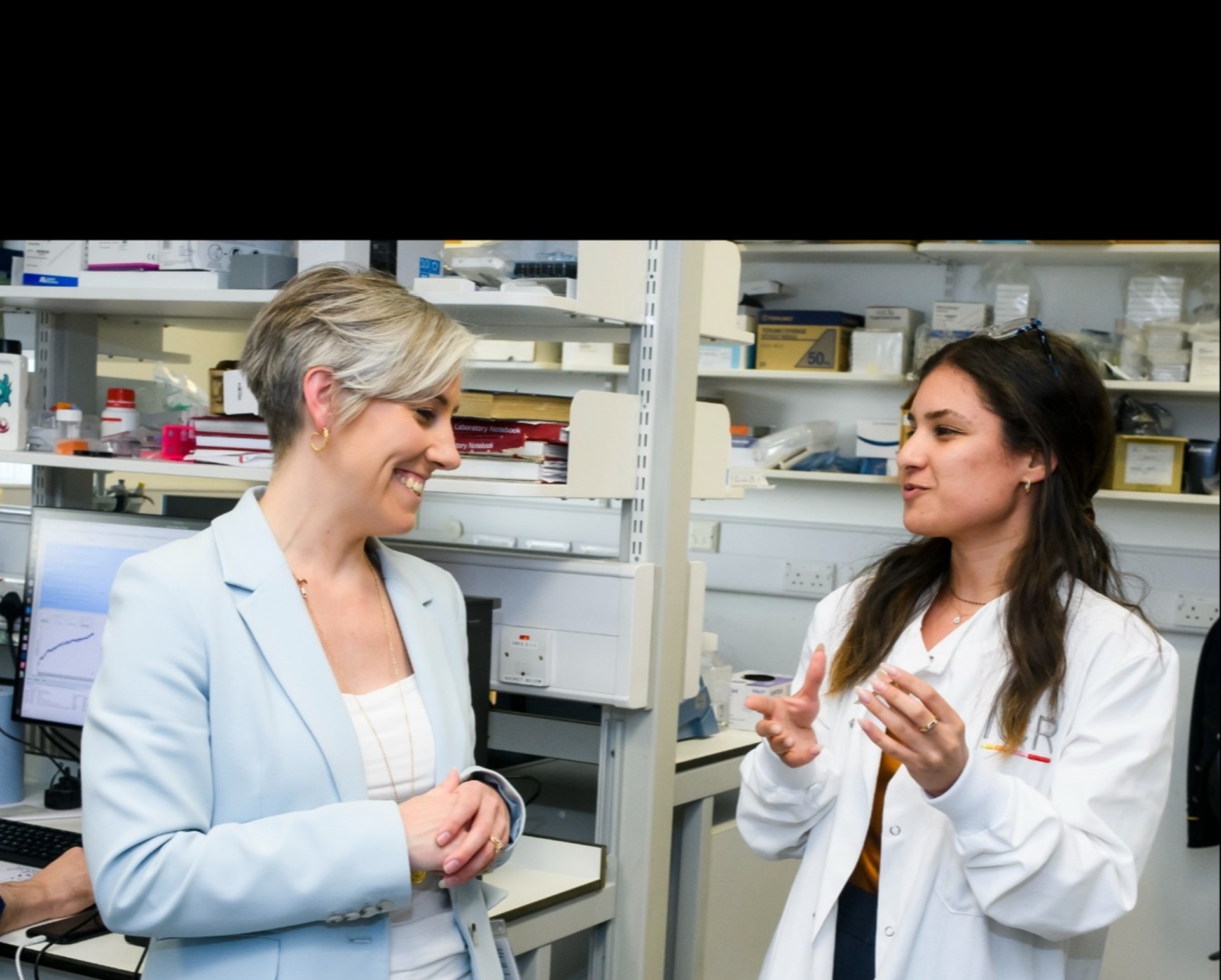 two women stand talking to one another in a Institute of Cancer Research laboratory, one wears a suit and another a lab coat