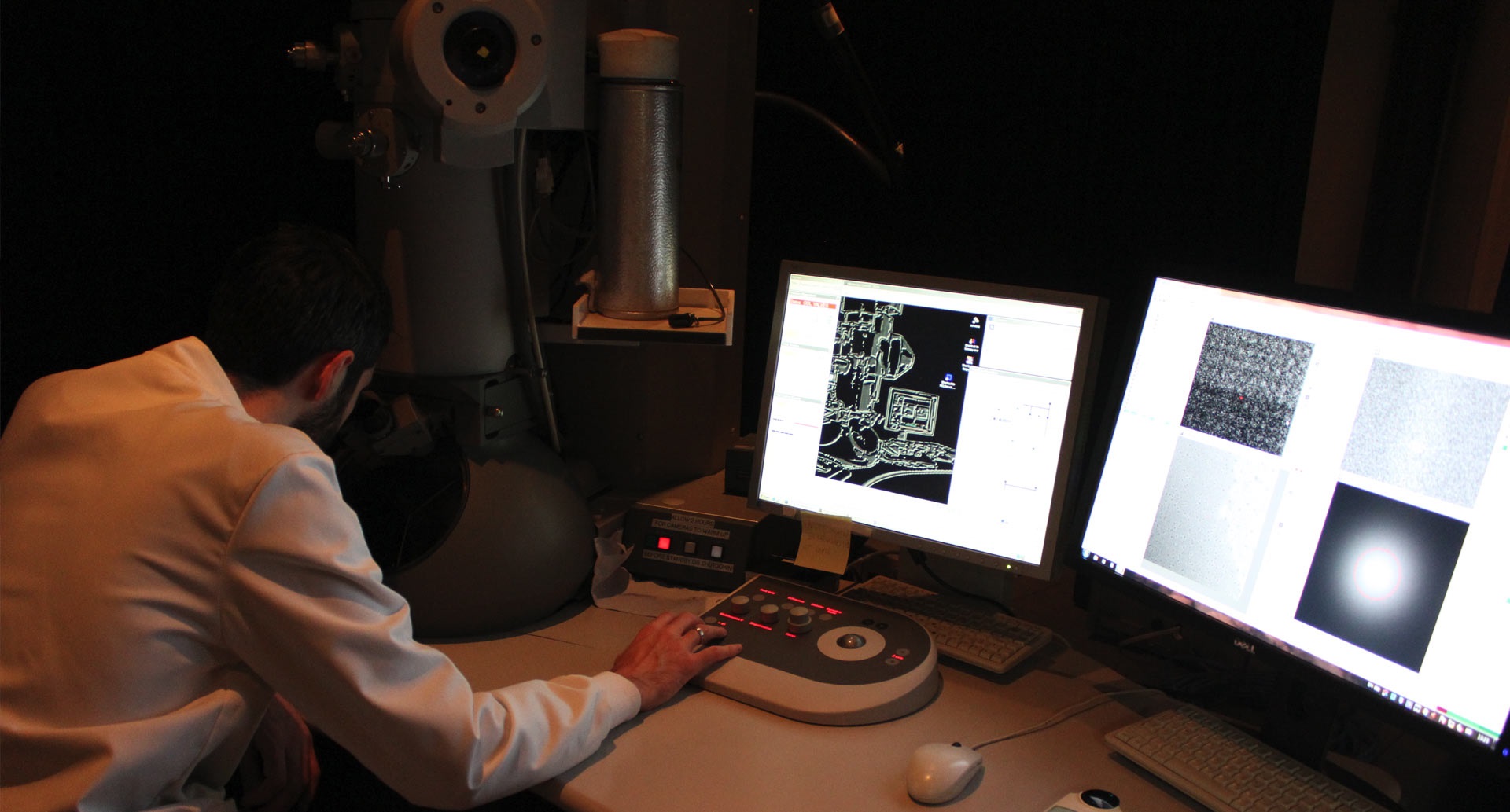 Researcher looking into a microscope, surrounded by several screens