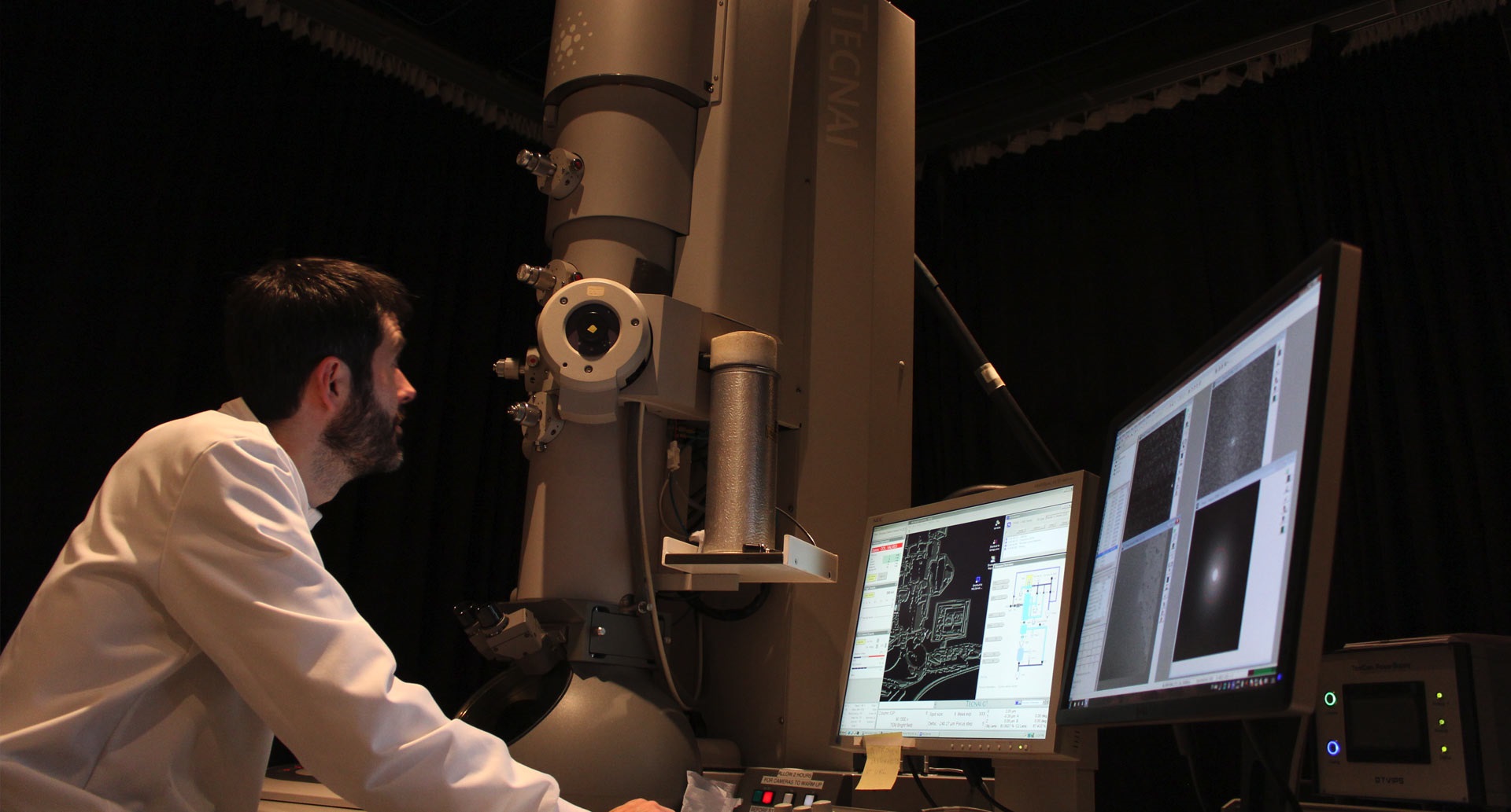 Researcher looking up at electron microscope, surrounded by several screens