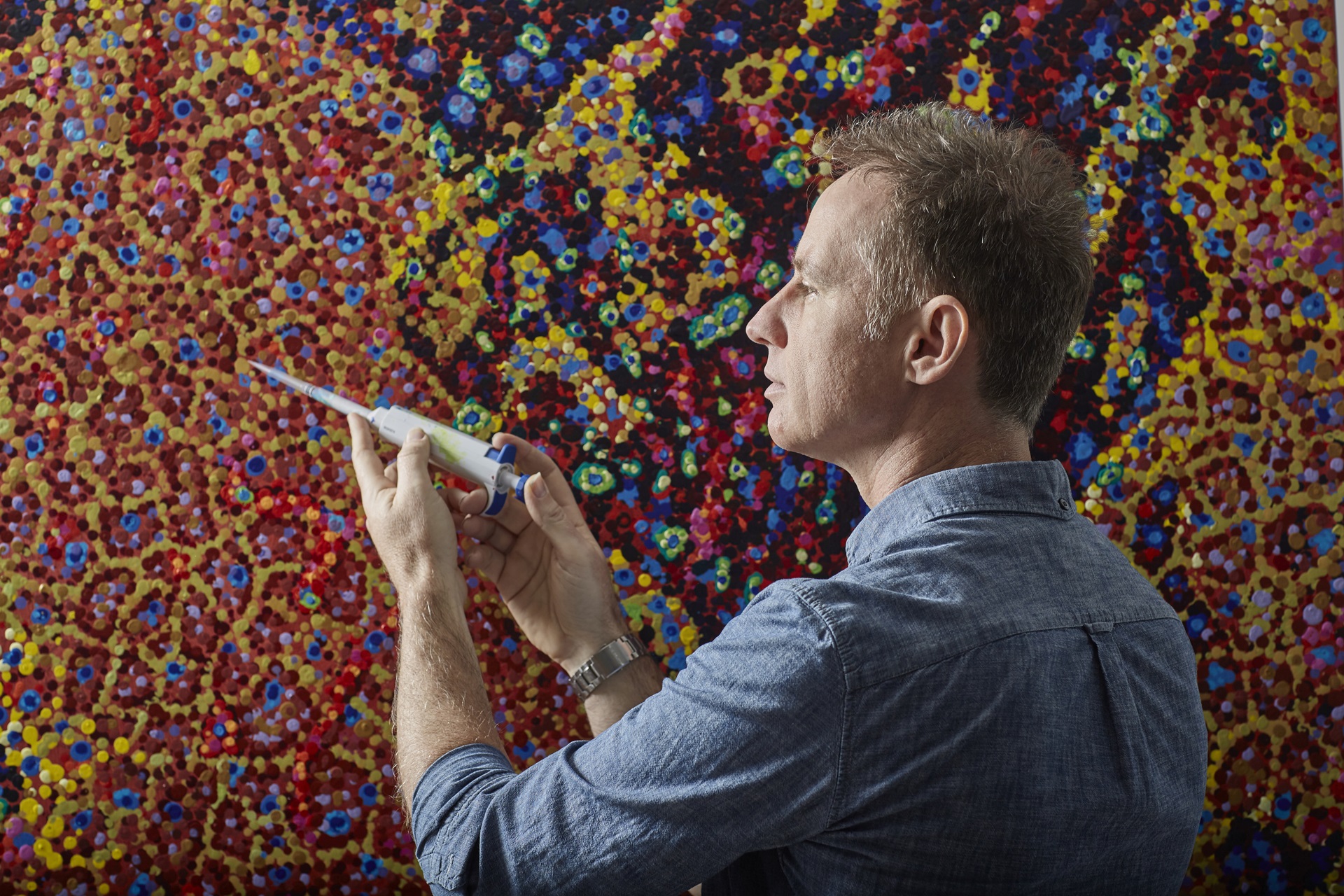 Philanthropy - pipette - a man stands in front of a painting of lots of colourful dots with a large pipette