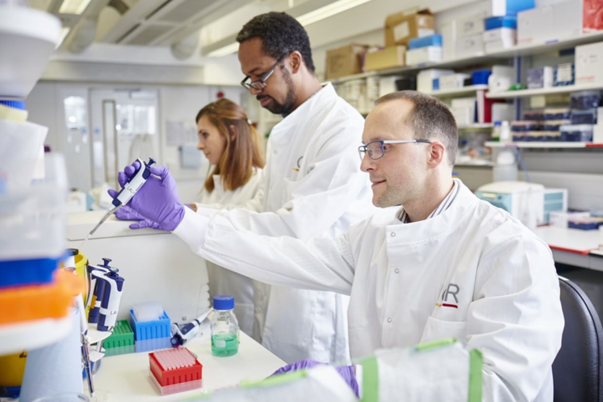 Three scientists in a lab conducting an experiment. One wears purple gloves.