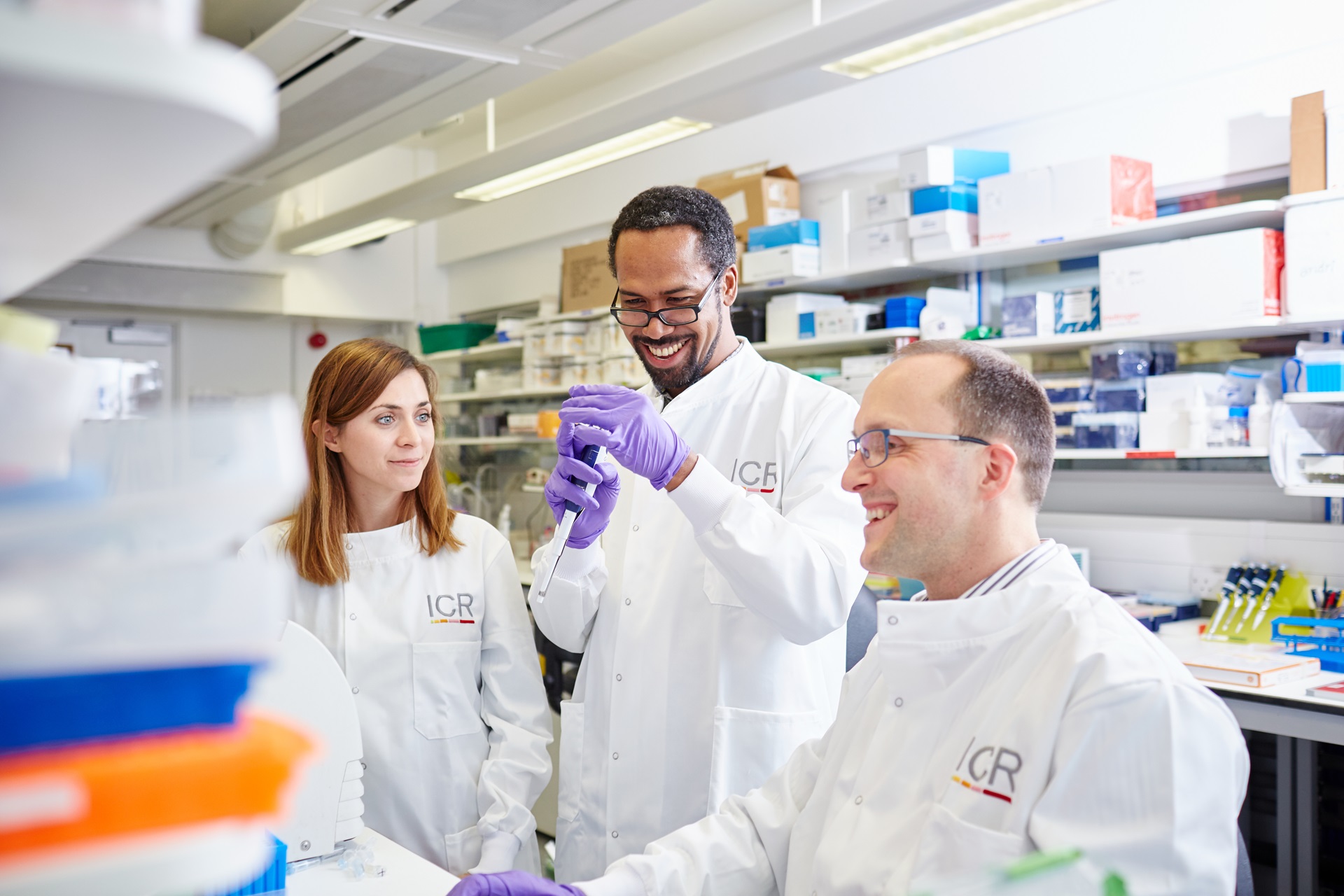 Three scientists in a lab conducting an experiment. One wears purple gloves.