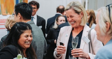 Three woman stand talking and laughing in a busy room. One holds a glass of champagne.