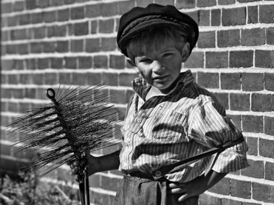 Black and white image of a young boy dressed in sooty clothes holding a chimney sweep