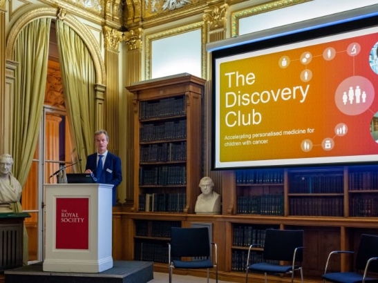 Speaker Kristian Helin at a podium in a plush venue giving a speech, with 'Discovery Club' on a projector behind him