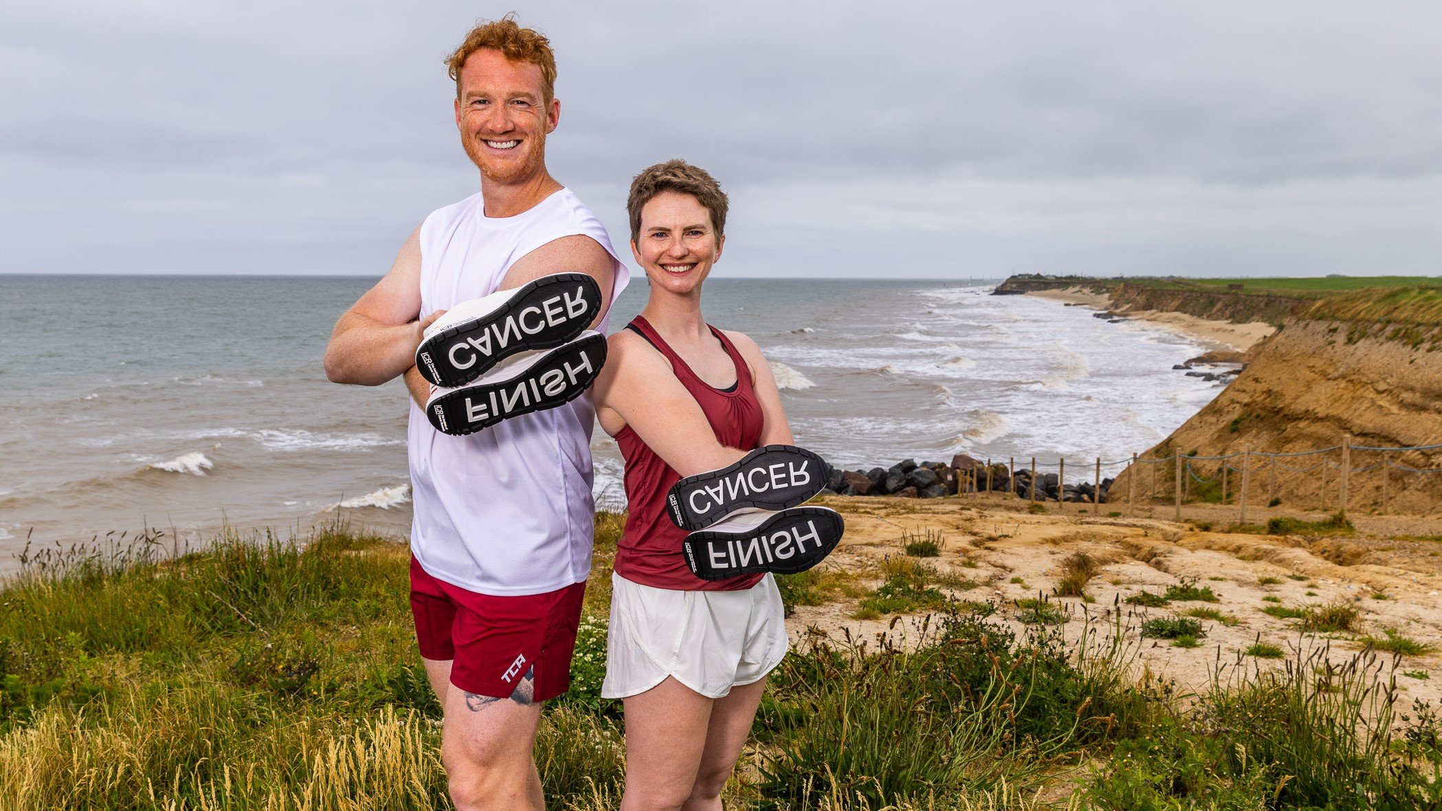 Sporting Champions Greg Rutherford (left) and Erin Kennedy (right) posing on a clifftop with the sea and beach behind them as they hold the custom trainers so you can see the words FINISH and CANCER on the soles.