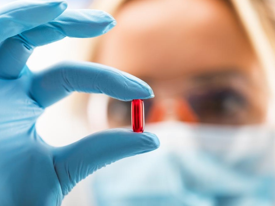 A scientist wearing a mask and blue gloves holds a red pill in focus