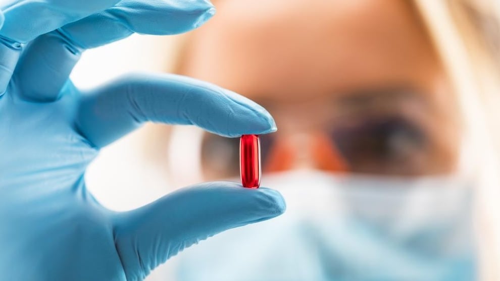 A scientist wearing a mask and blue gloves holds a red pill in focus.